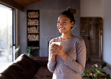 a smiling person with black curly hair in a bun holding a cup of hot chocolate in their living room using self-care tips to cope with holiday stress