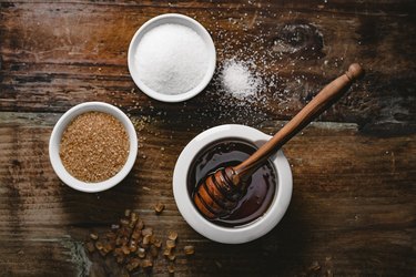 overhead view of three small bowls of white sugar, brown sugar and honey  on a dark wooden table