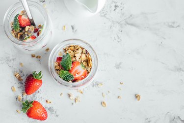 Two jars with tasty parfaits made of granola, strawberries and yogurt on white marble table. Top view, copy space